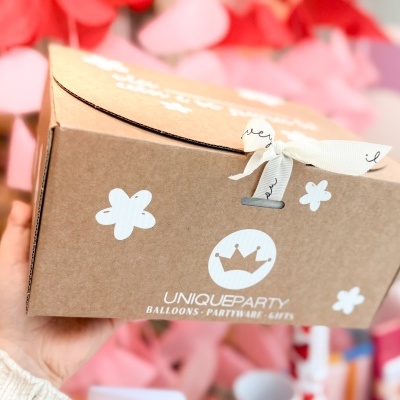 Caixa de cartão castanha UNIQUEPARTY com flores brancas e fita amarrada sobre fundo colorido.