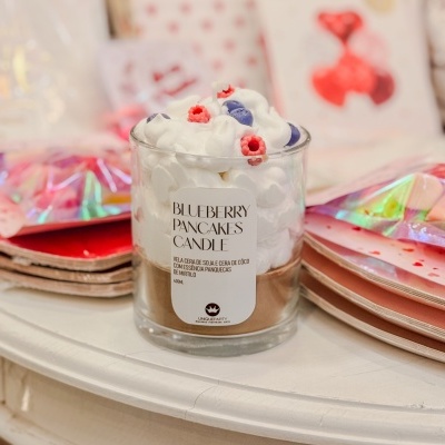 Vela em vidro decorada com cera imitando panquecas e chantilly sobre mesa branca com pratos e guardanapos coloridos