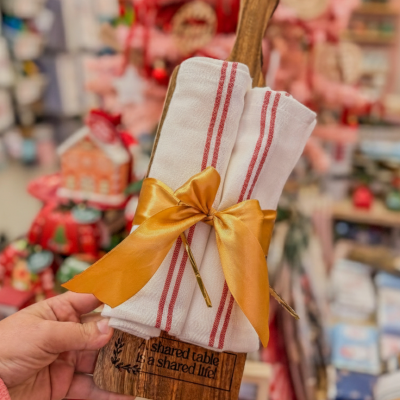 Tábua de madeira com panos de cozinha brancos com riscas vermelhas amarrados com laço dourado num ambiente de loja decorativa