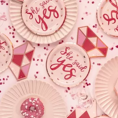 Pratos de papel rosa claro com texto She said YES, confetes e anéis decorativos em forma de diamante