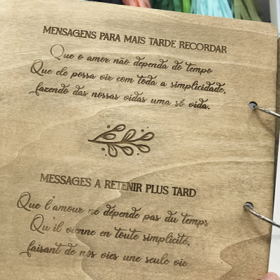 Livro artesanal em madeira com texto gravado em português e francês, decoração com galho, fundo com tecidos coloridos.