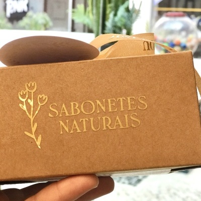 Caixa de sabonetes naturais castanha com texto dourado e flores, segurada por mão