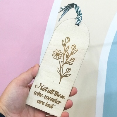 Marcador de livros em madeira gravado com flores e texto, com fita aos riscos, segurado por uma mão