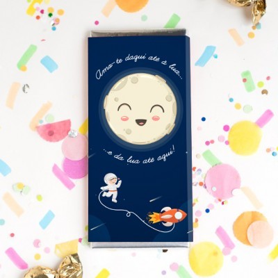 Embalagem de chocolate azul com lua sorridente, astronauta, foguetão e texto em português, sobre confetis coloridos