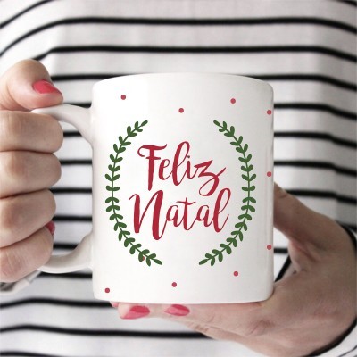 Caneca branca com texto Feliz Natal em vermelho e folhas verdes, segurada por mãos com unhas vermelhas