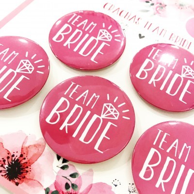 Cinco botons cor-de-rosa com texto branco 'TEAM BRIDE' e desenho de anel, sobre fundo branco floral.