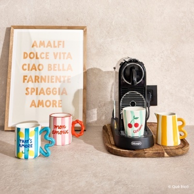 Máquina de café preta DeLonghi com chávenas e jarro coloridos em base de madeira e quadro decorativo com texto