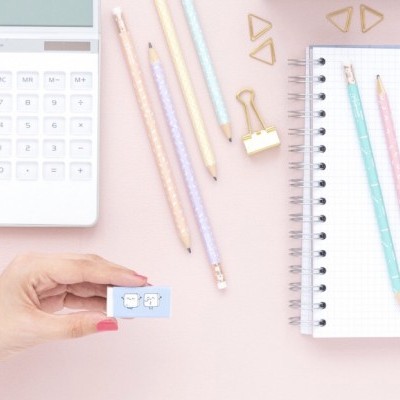 Mão segurando borracha azul clara com desenhos de nuvens e sol, lápis coloridos, caderno quadriculado, clips e calculadora num fundo rosa.