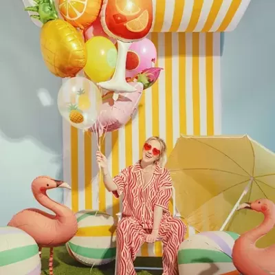 Mulher com pijama vermelho e branco segurando balões de frutas num cenário colorido com flamingos e bolas de praia.