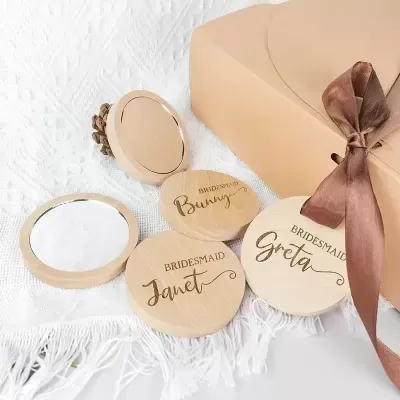 Espelhos compactos redondos em madeira clara com nomes em tinta dourada e caixa de presente bege com laço castanho