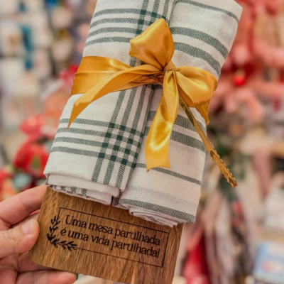 Panos de cozinha brancos com riscas verdes e fita dourada numa tábua de madeira gravada.