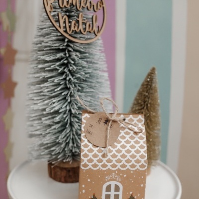 Caixa de papel castanha decorada para Natal com etiqueta, em mesa branca e árvores de Natal ao fundo