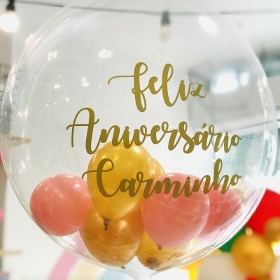 Balão transparente com texto dourado 'Feliz Aniversário Carminho' e balões interiores cor dourada e rosa