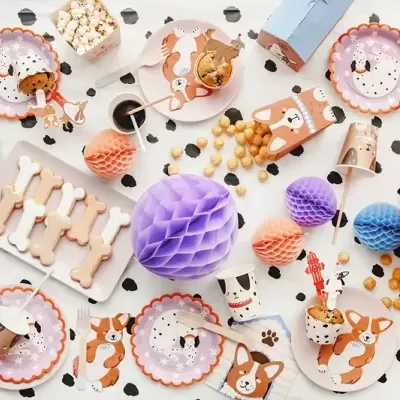 Utensílios de festa com tema de cães em tons rosa, azul e lilás sobre mesa branca manchada de preto