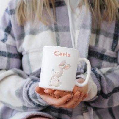 Caneca branca com coelho e nome Carla, sendo segurada por pessoa com casaco xadrez cinza e rosa