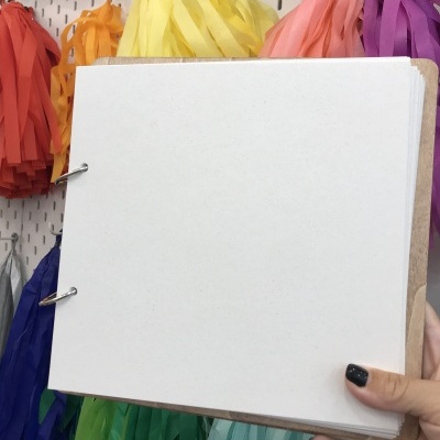 Caderno artesanal com capas de madeira e folhas brancas segurado à frente de decoração colorida em franjas de papel