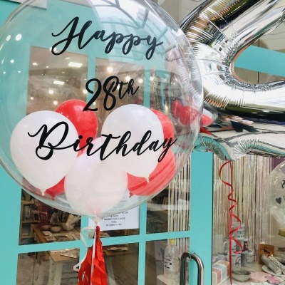 Balão transparente com texto 'Happy 28th Birthday' e balões internos vermelhos e brancos, junto a porta azul e balão prateado número.