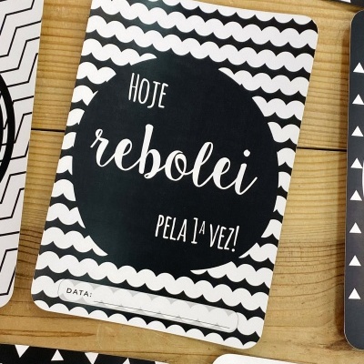 Cartão para bebé preto e branco com texto sobre primeira vez que rebolou, com espaço para data, sobre mesa de madeira.