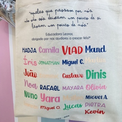 Saco de tecido bege com texto colorido e nomes imprimidos, pendurado num cabide prateado