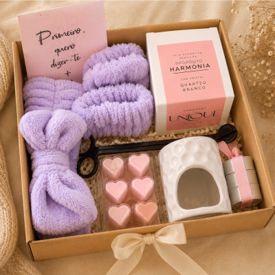 Caixa de presente com faixa creme em cetim contendo faixa de cabelo lilás, vela aromática, placas de cera em formato de coração, queimador branco e cartão rosa