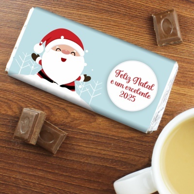 Barra de chocolate com embalagem azul clara e Pai Natal, com texto natalício, em mesa de madeira com chocolate e chávena branca.