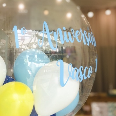 Balão transparente com balões coloridos e texto azul '1º Aniversário Vasco J'