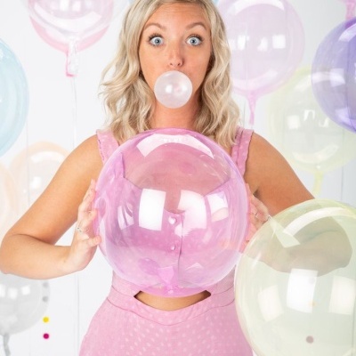 Mulher loira segurando balões transparentes coloridos e fazendo bolha com chiclete