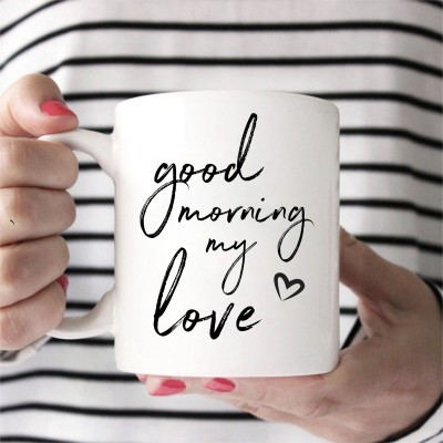 Caneca branca com texto 'good morning my love' em preto, segurada por mãos femininas com unhas rosas e camisa de riscas.