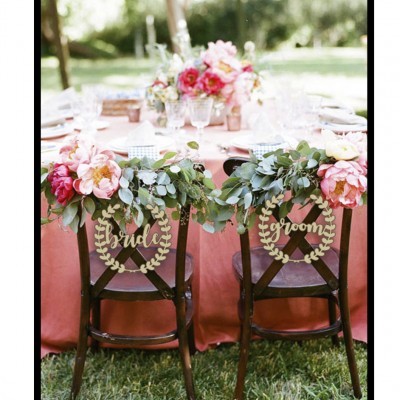 Cadeiras decoradas Bride e Groom com flores e folhas verdes à frente de mesa com toalha rosa ao ar livre.