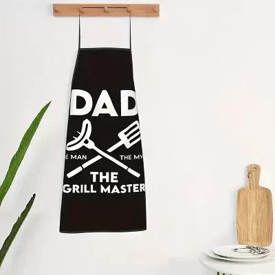 Avental preto com texto branco e desenhos de utensílios de grelha pendurado numa parede branca