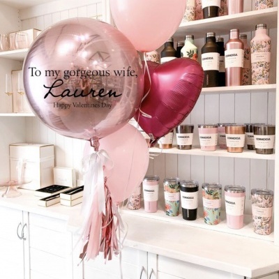 Balões decorativos em tons de rosa com texto visível em balcão branco com produtos decorativos em prateleiras