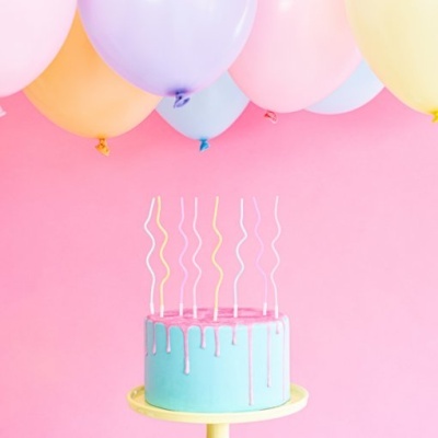 Bolo azul com cobertura rosa, velas onduladas e balões coloridos no fundo