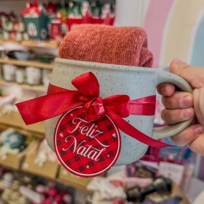 Caneca cinza com laço vermelho e etiqueta 'Feliz Natal' contendo tecido vermelho