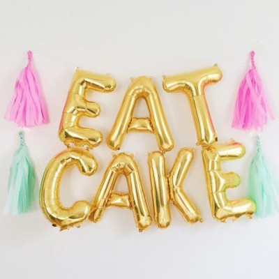 Balões dourados a dizer 'EAT CAKE' com pompons rosa e verde-água sobre fundo branco