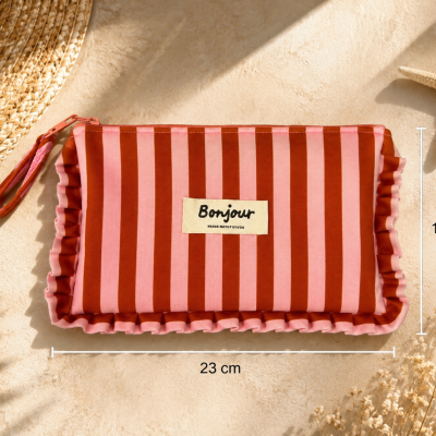 Necessaire listrada rosa e vermelha com etiqueta Bonjour sobre superfície bege com chapéu e decoração de praia.