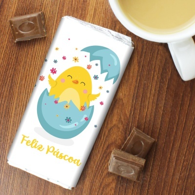 Barra de chocolate embrulhada com ilustração de pintainho e texto Feliz Páscoa sobre mesa de madeira com chocolate e chávena