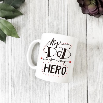 Caneca branca com texto 'My Dad is my Hero' sobre fundo de madeira clara com folhas e suculenta