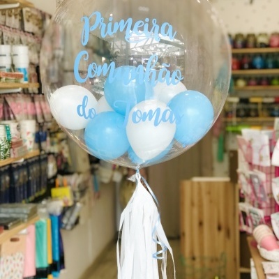 Balão transparente com texto azul e pequenos balões azuis e brancos dentro, pendurado em loja com prateleiras ao fundo