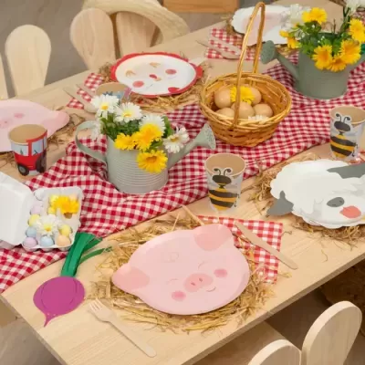 Mesa de madeira com decoração temática infantil com pratos de papel de animais, copos coloridos, flores em regadores, ovos em cesto, guardanapos xadrez e ovos de chocolate coloridos.