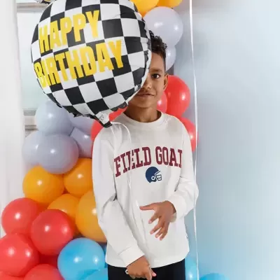 Menino com camisola branca segurando balão quadriculado 'HAPPY BIRTHDAY' e balões coloridos ao fundo