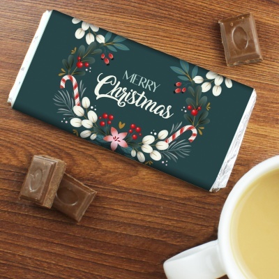 Barra de chocolate natalícia com decoração verde e texto Merry Christmas, com pedaços de chocolate e chávena branca numa mesa de madeira