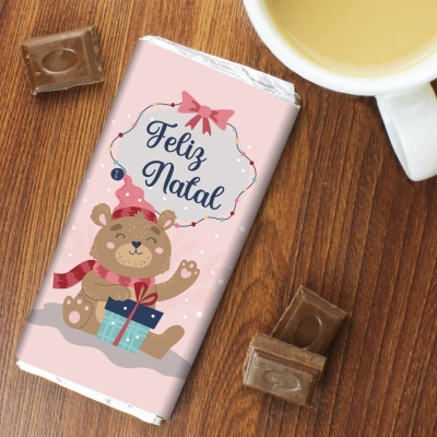 Barra de chocolate de embalagem rosa com ursinho e texto Feliz Natal junto a pedaços de chocolate e chávena
