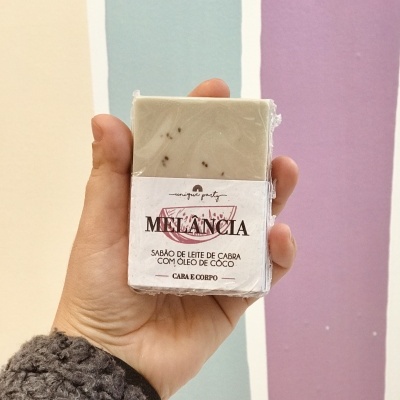 Sabão MELANCIA com leite de cabra e óleo de coco segurado por mão com fundo colorido