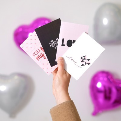 Mão segura cartões coloridos com mensagens de amor e corações em fundo desfocado com balões em forma de coração.