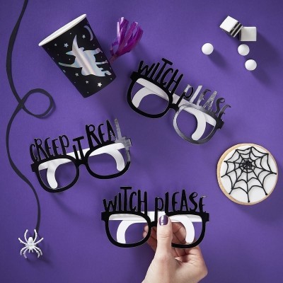Óculos pretos com frases de Halloween, copo com morcego, bolacha decorada e aranha de plástico sobre fundo roxo.