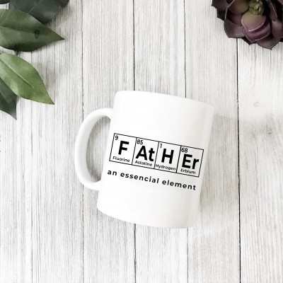 Caneca branca com design de tabela periódica e texto 'FATHER an essencial element' sobre mesa de madeira clara