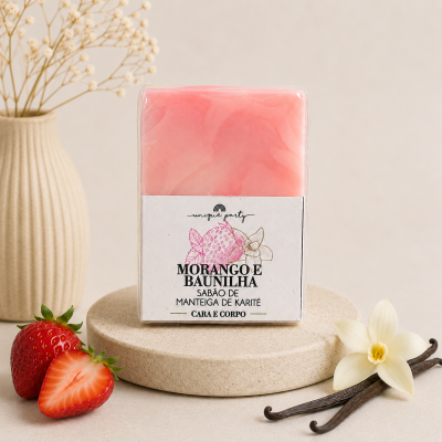 Bloco de sabão rosa embalado com rótulo branco com texto Morango e Baunilha colocado em base redonda creme com morangos, flor branca e vagem de baunilha à volta