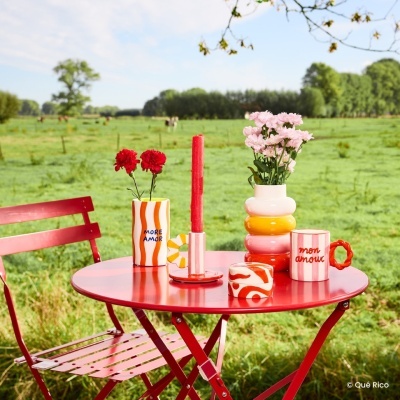 Mesa e cadeiras vermelhas com vasos cerâmicos coloridos decorados com flores ao ar livre num campo verde