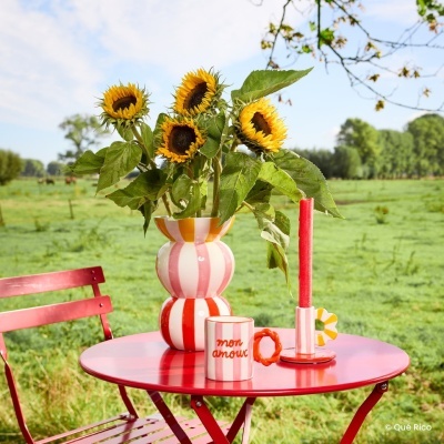 Mesa e cadeira vermelhas em metal com vaso, girassóis, vela e caneca com texto em cenário campestre.