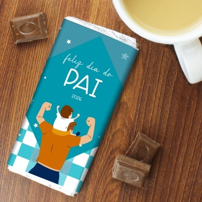 Barra de chocolate com embalagem azul para o Dia do Pai, com desenho de pai e filho, sobre mesa de madeira com chocolate e chá